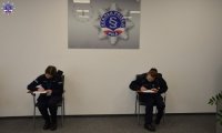 Dwie policjantki wypełniają dokumenty siedząc na krześle. W tle wiszący na ścianie zegar z logo Szkoły Policji w Pile.
